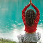 A woman meditates in nature on a rock by a tranquil turquoise lake, practicing yoga.